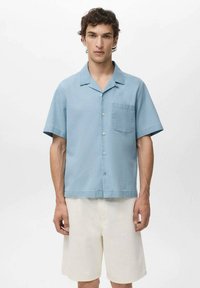 Camisa de manga corta en azul claro, hecha de una tela de textura suave, con un bolsillo en el pecho y un diseño abotonado. Combinada con pantalones cortos color crema.