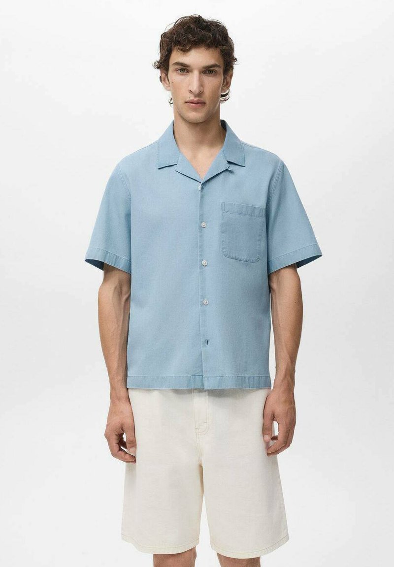 Camisa de manga corta en azul claro, hecha de una tela de textura suave, con un bolsillo en el pecho y un diseño abotonado. Combinada con pantalones cortos color crema.