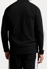 Černá mikina s vysokým límcem, raglánovými rukávy a bílými pruhy po stranách. Tkanina působí hladce a pružně, lem je žebrovaný.