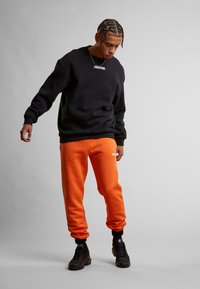 Sweatshirt noir avec le logo "STARTER" blanc, associé à un pantalon de jogging orange vif. Baskets noires, bas de pantalon ajustés. Texture lisse.