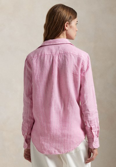 Kvinde set fra siden iført en lyserød og hvid langærmet skjorte med vertikale striber og hvide bukser mod en ensfarvet baggrund.