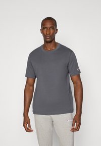 Graues Baumwoll-T-Shirt mit Rundhalsausschnitt und kurzen Ärmeln, ausgestattet mit einem kleinen Logo auf dem linken Ärmel, kombiniert mit hellgrauen Jogginghosen.