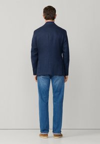 Mann mit dem Rücken zur Kamera trägt ein marineblau kariertes Sakko, hellblaue Jeans und beige Freizeitschuhe vor einem einfarbigen hellen Hintergrund.