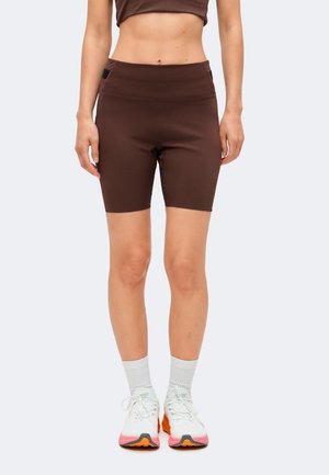 Kvinde iført brune high-waist biker shorts, hvide sokker og hvide løbesko med pink og orange såler, står mod en ensfarvet baggrund.