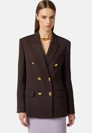 Donna che indossa un blazer doppiopetto marrone scuro con bottoni dorati e una collana choker dorata, in piedi davanti a uno sfondo semplice.