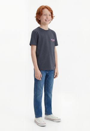 Punapea poiss naeratab, kandes tumedat halli trükiga t-särki, siniseid pükse ja valgeid tossusid, seistes valge tausta ees.