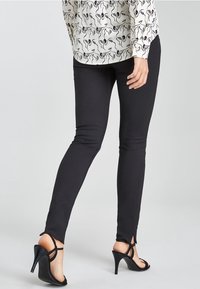 Pantalons skinny noirs avec une texture lisse, présentant des fentes latérales au niveau des chevilles. Associés à des sandales noires à talons hauts avec des lanières.