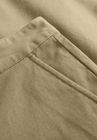Tissu beige avec une surface lisse et texturée ; présente des coutures cousues et une poche latérale, mettant en valeur des lignes épurées et un design simple.