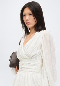 Γυναίκα με μακριά σκουρόχρωμα μαλλιά, φορώντας ένα λευκό ρουζάτο φόρεμα με υφή, κρατά μια μικρή σκουρόχρωμη τσάντα στον ώμο της, κοιτάζοντας στο πλάι.