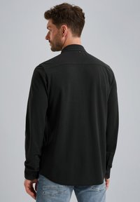 Camisa negra de manga larga con una textura suave, cuello clásico y acentos de botones, mostrada desde la parte de atrás sobre un fondo gris claro.