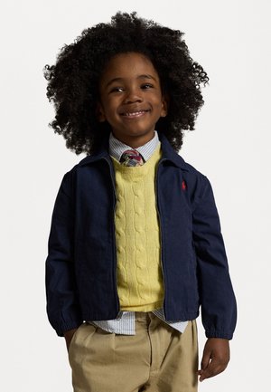 Navy blue zip-up jacket, yellow cable-knit sweater, white and blue striped shirt, khaki pants, with a red logo detail on the jacket.