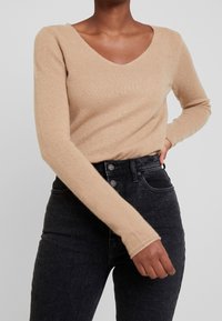 Maglione beige a maniche lunghe con scollo a V, realizzato in morbido tessuto a maglia; indossato con jeans neri a vita alta.