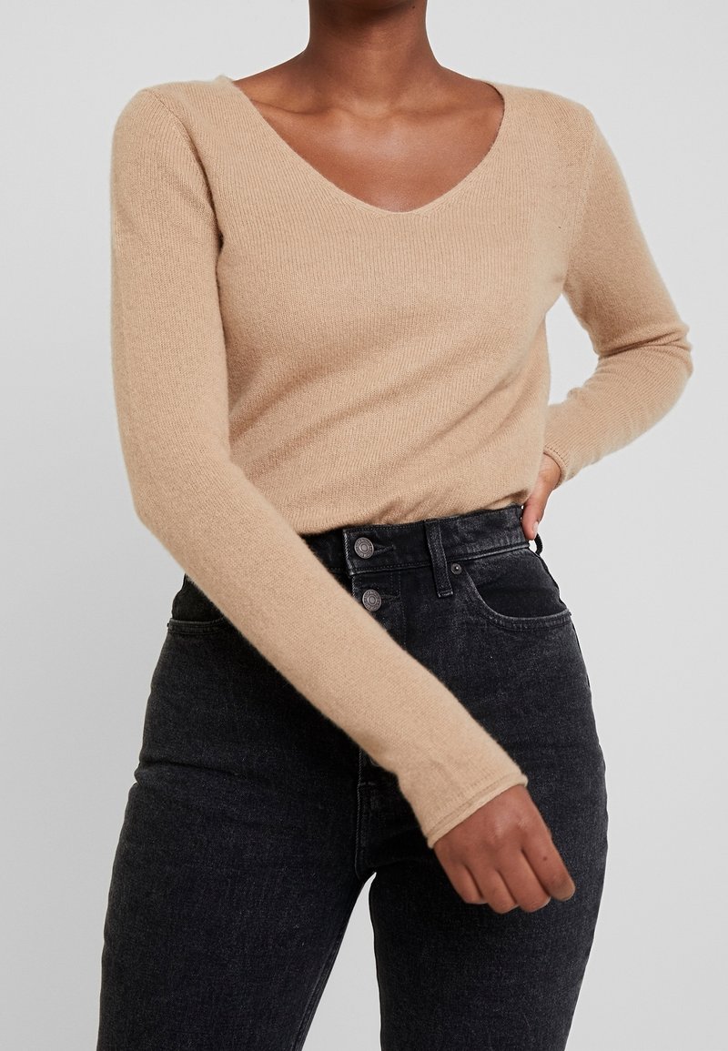 Maglione beige a maniche lunghe con scollo a V, realizzato in morbido tessuto a maglia; indossato con jeans neri a vita alta.