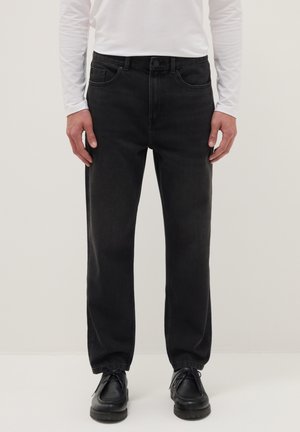 Homme portant un jean noir à jambes droites, des chaussures noires à lacets, et une chemise blanche à manches longues, sur un fond uni.