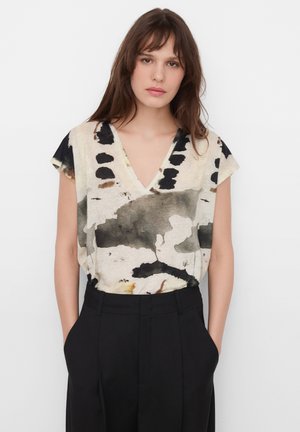 Frau mit braunen Haaren trägt ein V-Ausschnitt-Top mit abstraktem Muster in Schwarz, Beige und Braun, in eine hochgeschnittene schwarze Hose gesteckt.