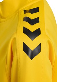 Gele stof met een zwart chevronpatroon dat verticaal is gedrukt op de mouw van een kledingstuk.