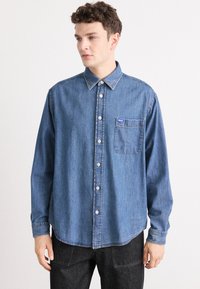 Camicia in denim blu con chiusura a bottoni, colletto a punta, taschino sul petto e maniche lunghe. Texture leggermente sbiadita, vestibilità dritta e cuciture a contrasto.