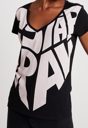 Personne portant un T-shirt noir à col en V avec de grandes lettres blanches en bloc formant le mot "STAR DAY" sur le devant.