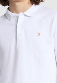 Polo blanc à manches longues en tissu texturé avec un col classique, doté d'une patte de boutonnage à trois boutons et d'un petit logo brodé en orange.