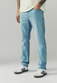 Blugi de denim bleu deschis cu croi drept, având cinci buzunare și un ușor efect de uzură, asortați cu adidași albi și dungi bleu închis.