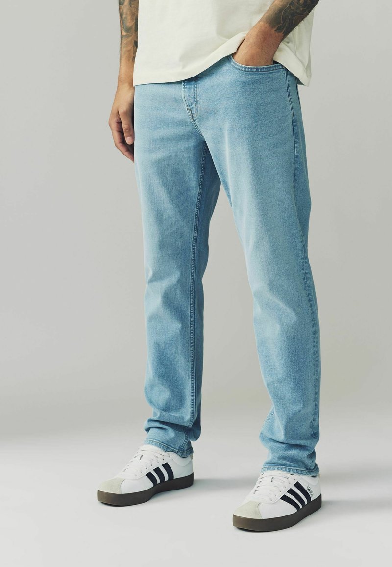 Blugi de denim bleu deschis cu croi drept, având cinci buzunare și un ușor efect de uzură, asortați cu adidași albi și dungi bleu închis.