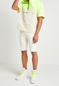 Lekki, krótki pulower z krótkim rękawem w neonowej zieleni i beżu, z zamykaną na zamek kieszenią, w połączeniu z off-white szortami dżinsowymi i białymi sneakersami.