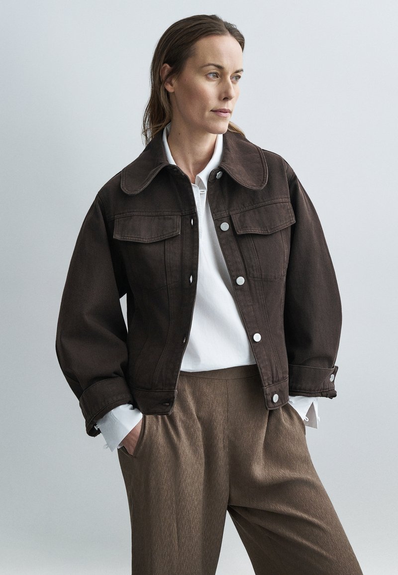 Ruda džinsinė striukė su dideliu apykakliu, sagų užsegimu ir priekinėmis kišenėmis, derinama su balta marškinėliais ir tekstūruotomis rudomis kelnėmis.