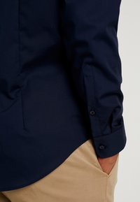 Granatowa koszula z guzikami i długimi rękawami, o gładkiej fakturze, z zaokrąglonym dołem i standardowymi mankietami, w połączeniu z spodnimi w kolorze khaki.