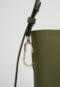 Záběr zblízka na olivově zelenou koženou tašku s nastavitelným popruhem a zlatým kovovým karabinkovým uzávěrem připevněným na boku.