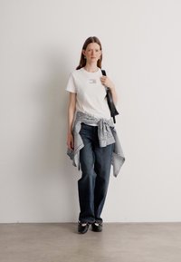 Hvid t-shirt med et lille grafik, blå stribet bælte omkring taljen, mørkeblå jeans og sorte slip-on sko. Minimalistisk design.
