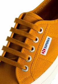 Sneaker in tela gialla con sei lacci marroni, occhielli argentati e una patch con logo rosso e blu sul lato. Suola in gomma bianca.