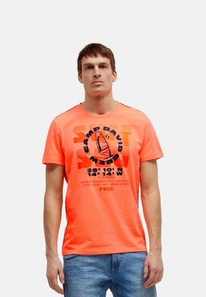 Mann trägt ein leuchtend oranges T-Shirt mit „Camp David Crew“-Schriftzug und Segelboot-Grafik, kombiniert mit blauen Jeans, steht vor weißem Hintergrund.
