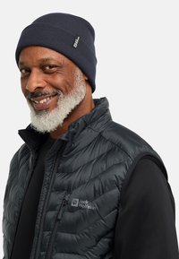 Uomo sorridente con barba grigia, che indossa un berretto di maglia scuro e un gilet trapuntato Jack Wolfskin nero sopra una maglietta a maniche lunghe nera.