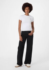 Hvid t-shirt med korte ærmer og et diskret rødt logo på brystet, kombineret med sorte bukser med vide ben og hvide sneakers.