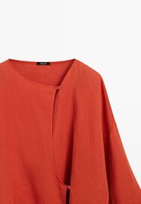 Rust-orange Leinenbluse mit rundem Ausschnitt, asymmetrischem Frontverschluss und zwei kleinen passenden Knöpfen.
