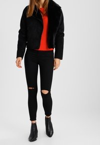 Czarna kurtka z futra sztucznego z zamkiem błyskawicznym, czerwona sweter z dzianiny, czarne jeansy skinny z przetarciami oraz czarne botki za kostkę. Prosty, nowoczesny design.