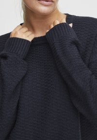 Pull en tricot bleu marine avec un motif texturé, des poignets côtelés et une coupe légèrement ample. Le tissu semble doux et chaleureux.
