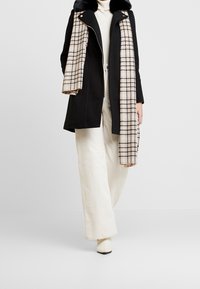 Abrigo de lana negro con cuello de piel, pantalones crema de canalé, y una bufanda de cuadros beige drapeada encima. El calzado es crema y tiene un ligero tacón.