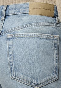 Ανοιχτό μπλε τζιν τσέπη πίσω με ορατή ραφή και καφέ δερμάτινη ετικέτα της μάρκας με την επιγραφή "SAMSOE SAMSOE" στη μέση.