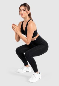 Vrouw in zwarte sportkleding en witte sneakers die een squat oefening doet tegen een effen lichte achtergrond.