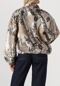 Bomberjack in een metallic zilver met een bloemmotief, voorzien van gouden en zwarte accenten; heeft een geplooide zoom en volumineuze mouwen.