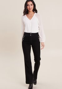 Pantalons noirs taille haute avec des boutons argentés et des accents latéraux, associés à un chemisier blanc avec des manches texturées à pois.