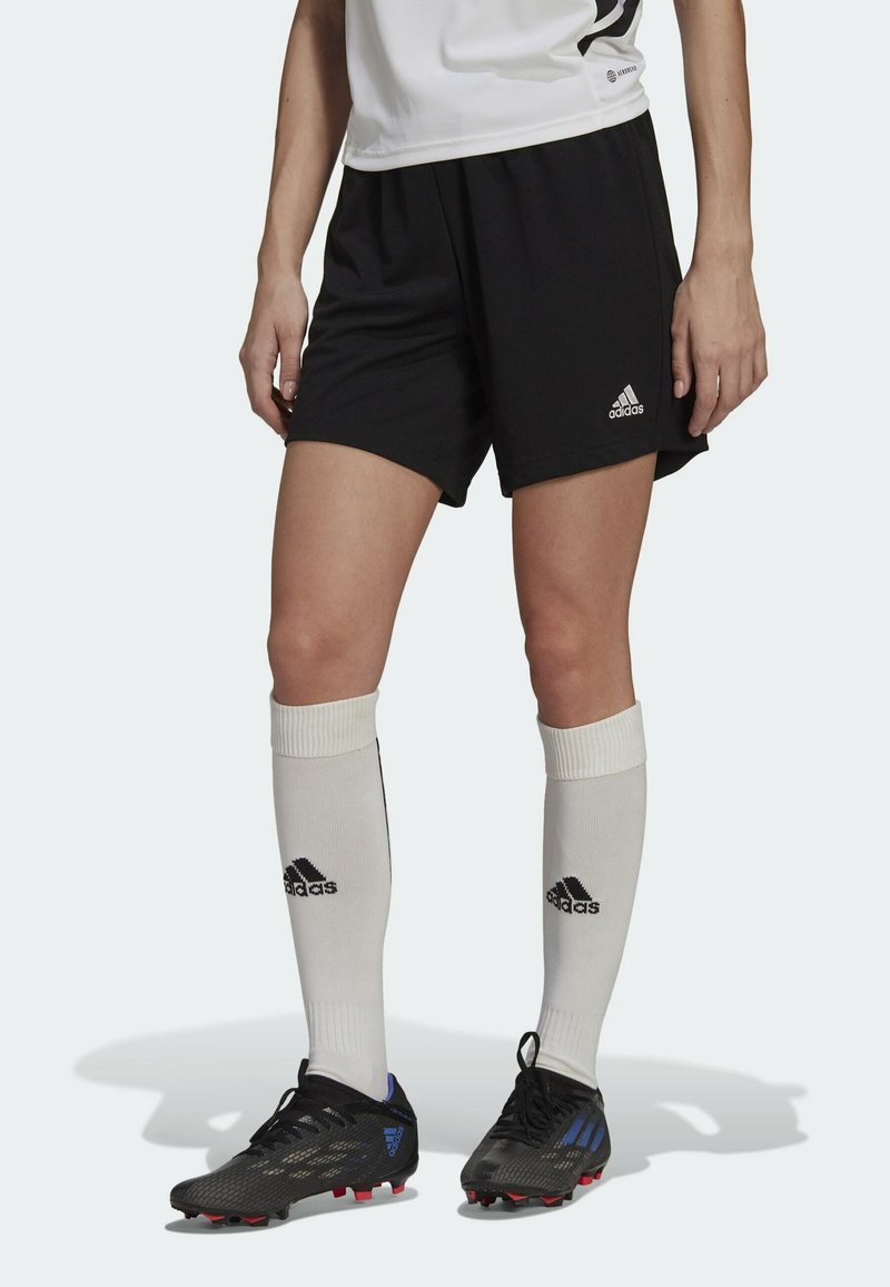 Personne portant un short noir Adidas, des chaussettes blanches Adidas montant jusqu'aux genoux et des crampons noirs, debout devant un fond clair et uni.