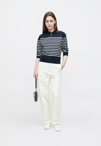 Pull-over rayé bleu marine et blanc avec un col, associé à un pantalon blanc taille haute et des baskets blanches. Le modèle tient un petit sac à main.
