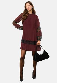 Rochie plisată burgund cu mâneci transparente din dantelă neagră și tiv din dantelă neagră, asortată cu o geantă neagră și pantofi cu toc înalt.