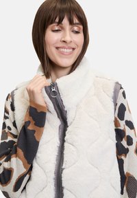 Faux-Fur-Weste in Creme mit gestepptem Design; gestrickte Ärmel mit einem braun-schwarzen Leopardenmuster; Reißverschluss vorne.