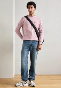 Gaiši rozā džemperis ar apaļu apkakli, zili džinsi un balti sneakers ar melniem akcentiem. Modelis stāv ar melnu crossbody somu.