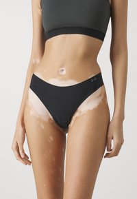 Gli slip bikini neri presentano un design a vita bassa, una trama liscia e un accento con logo sul lato, abbinati a tonalità e texture della pelle naturale.