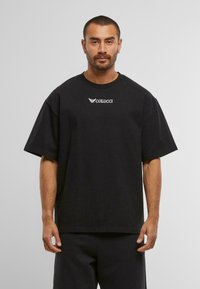 Mann med kort hår og øredobber iført oversized svart Colucci t-skjorte og svarte bukser, stående mot en enkel grå bakgrunn.