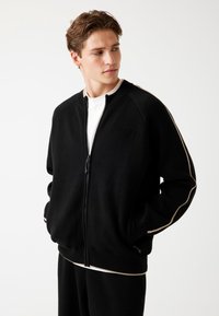 Schwarze Kapuzenjacke mit hohem Kragen, mit dezentem Logo und kontrastierenden Beigen Streifen entlang der Ärmel. Weicher Stoff und lockere Passform.
